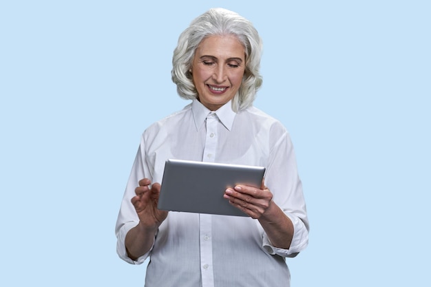 Smiling senior grayhaired woman using tablet pc isolated on pale blue background