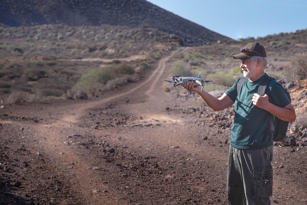 Smiling senior man holding a drone in his hand enjoying excursion in outdoor Arid landscape in background