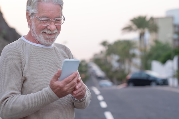 Smiling senior man standing in the street looking at mobile phone enjoying tech and social