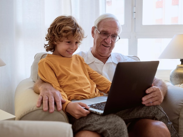 Smiling senior man with grandson watching interesting video on laptop while sitting together in armchair in living room at home