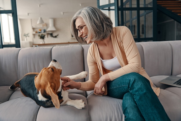 Smiling senior woman in casual clothing spending time with her dog while sitting on the sofa at home