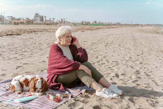 Smiling senior woman sitting on the beach beside her sleeping dog during the phone conversation