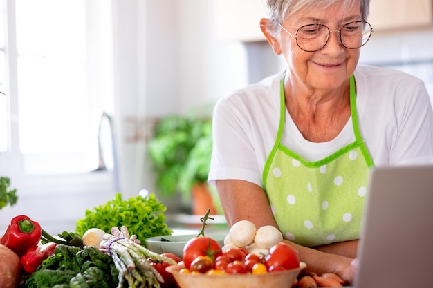 Smiling senior woman surfing the net sitting in the home kitchen while preparing vegetables Mature woman using laptop technology looking for new recipes