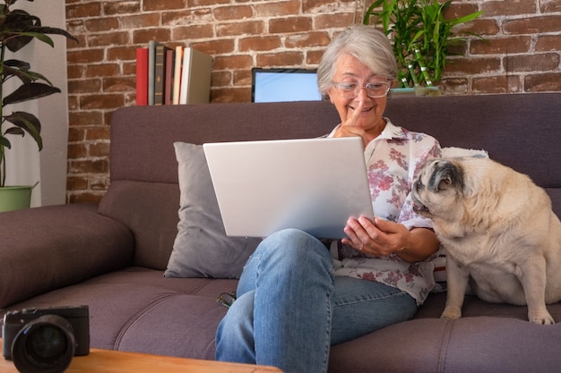 Smiling senior woman talking to her pug dog while sitting on sofa at home using laptop computer