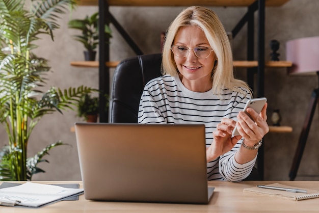 Smiling senior woman using smart phone and laptop at home office