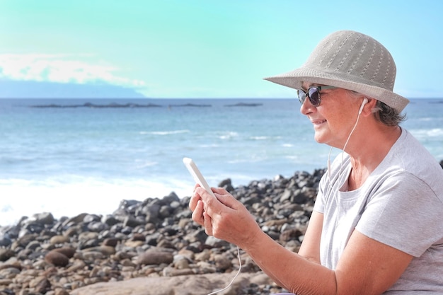 Smiling senior woman wearing hat and sunglasses sitting on the beach using mobile phone sending message Holding telephone with white cover Horizon over water