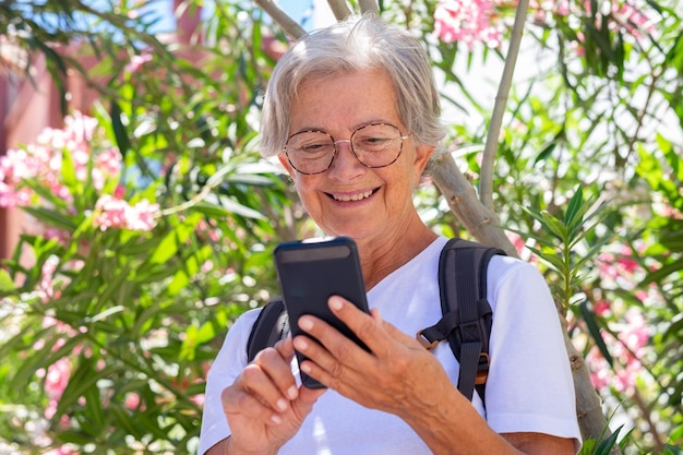 Smiling senior woman with eyeglasses holding mobile phone while standing under a flowered plant summer sunny day