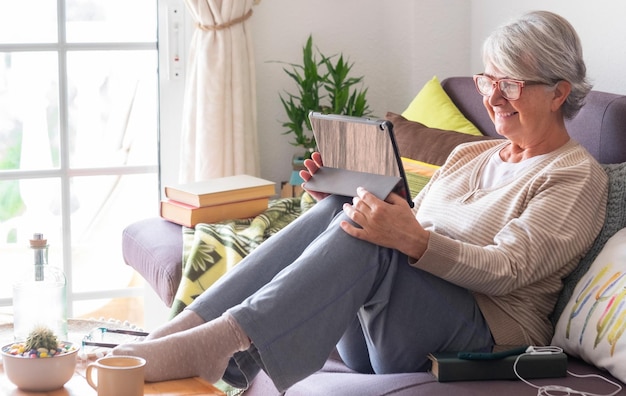 smiling senior woman with white hair relaxes at home lying on the sofa looking at her digital tablet