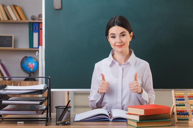 smiling showing thumbs up young female teacher sitting at desk with school tools in classroom