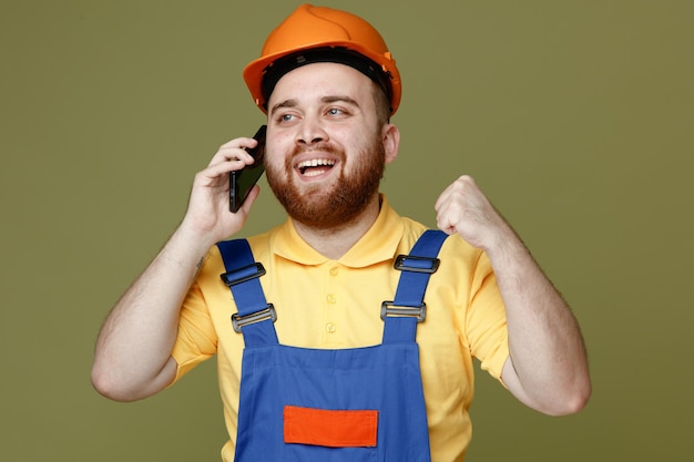 Smiling showing yes gesture speaks on phone young builder man in uniform isolated on green background