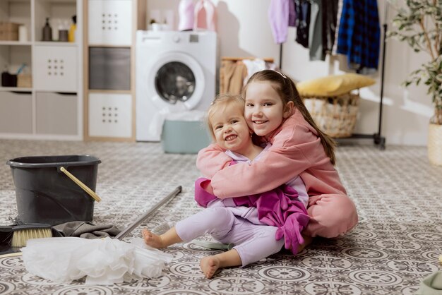 Smiling sisters sit on laundry room floor hugging cooperation while cleaning with rubber gloves sibling love shared household chores