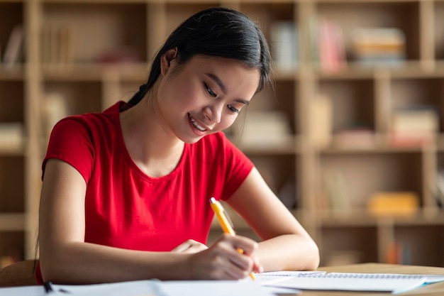 Smiling smart young chinese woman student doing homework preparing to exam alone in room interior close up