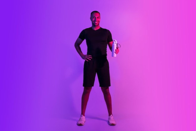 Smiling sporty black guy holds bottle of water purple background