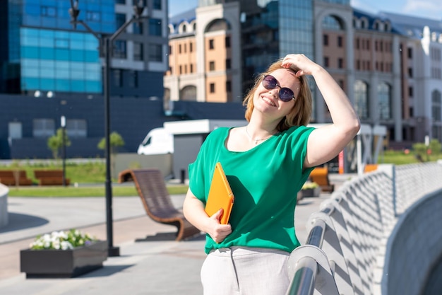 A smiling student girl in sunglasses holding a notebook or tablet in her hands on the embankment on the street