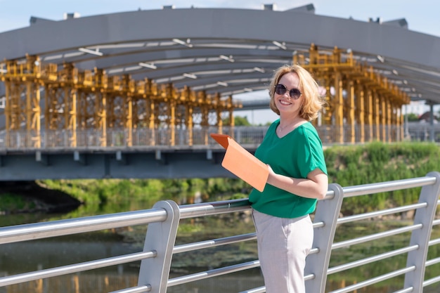 A smiling student girl in sunglasses holding a tablet in her hands reading news using applications on the street