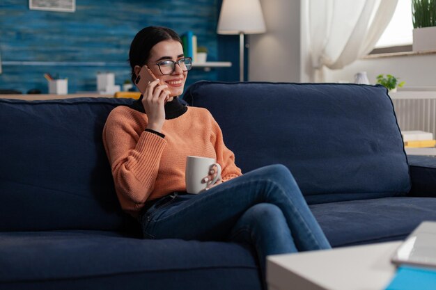 Smiling student sitting on sofa talking on smartphone. College girl with glasses holding cup having casual phone call. Freelancer on couch having happy phonecall with friend on mobile phone.