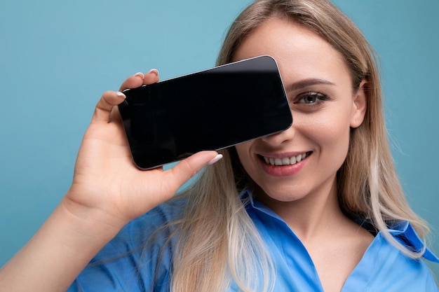 Smiling stylish blond girl holding smartphone screen with mockup for web page near her face on blue