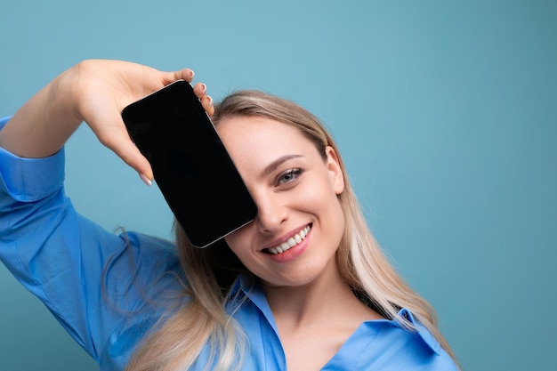 Smiling stylish blond girl holding smartphone screen with mockup for web page near her face on blue