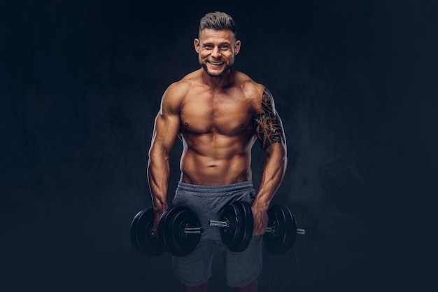 Smiling stylish bodybuilder with a tattoo on his arm, doing the exercises with dumbbells. Isolated on a dark background.