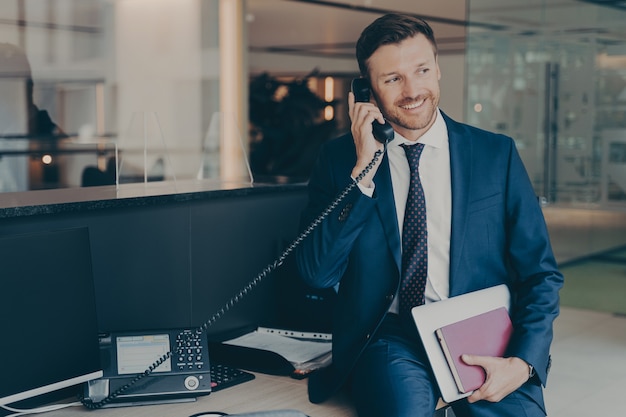 Smiling successful businessman in suit talking on phone sitting in office on top of desk, holding laptop and agenda, answering office call, checking implementation of recommendations and daily reports