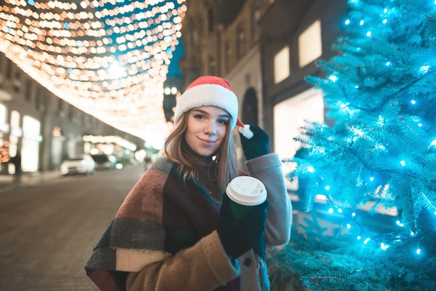 Smiling sweet girl in a Christmas hat and a cup of coffee in her hands stands at the Christmas tree on the street