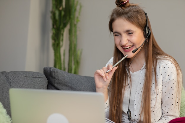 Smiling teen girl wearing headphones listening to audio course using laptop at home, making notes, young woman learning foreign languages, digital self education, studying online