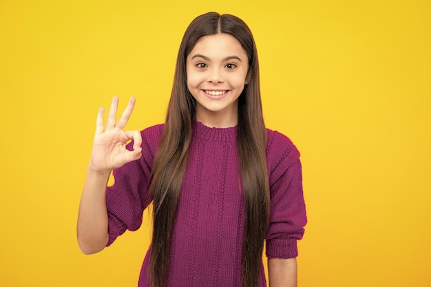 Smiling teen girl young teenager showing ok hand sign looking at camera isolated on yellow studio background