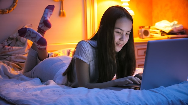 Smiling teenage girl lying on bed at night and chatting on laptop.