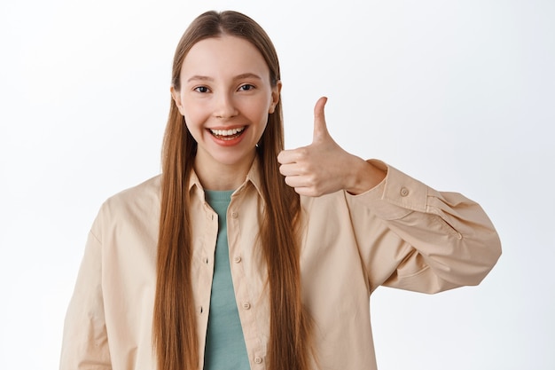 Smiling teenage girl shows thumbs up and say yes, nod in approval, praise excellent choice, recommends something cool, standing pleased against white wall