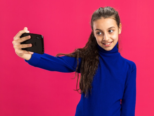 Smiling teenage girl taking selfie isolated on pink wall