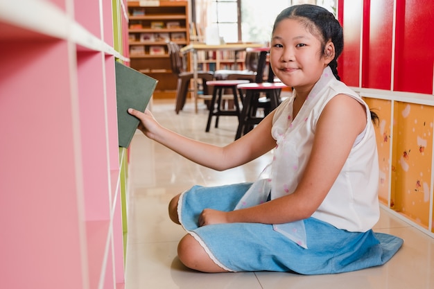 Smiling teenager girl student choosing books from shelf library in school.