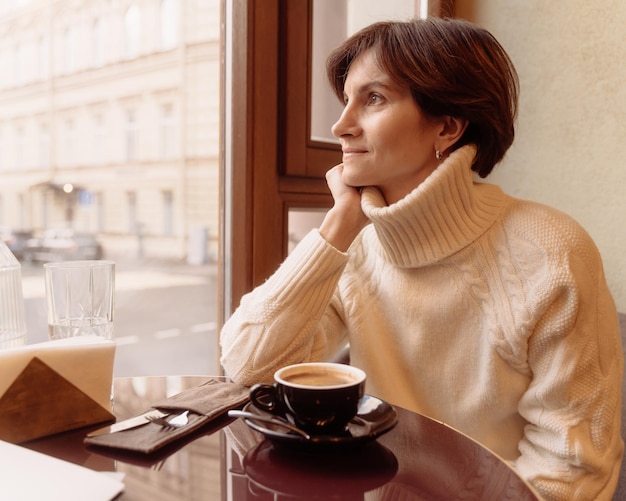 Smiling and thoughtful woman in sweater looking at window in cafe while drinking coffee