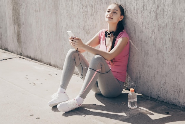 Smiling tired girl sitting on the ground outdoors resting after a hard workout and drinking water