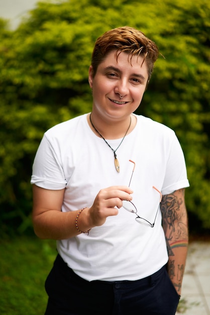 Photo smiling transgender man outdoors in casual attire confidence evident in stance with glasses in hand tattoo visible on arm plants backdrop suggesting fresh start inclusivity gender identity pride