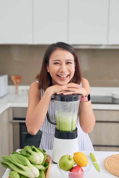 Smiling vegan Asian woman making a smoothie with fruit and vegetable in the kitchen