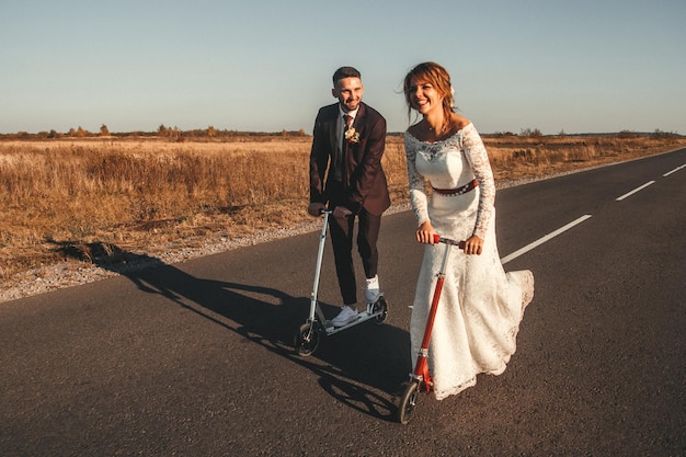 Smiling wedding couple riding a on scooters along the road outside the city at sunset