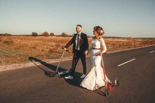 Smiling wedding couple riding a on scooters along the road outside the city at sunset