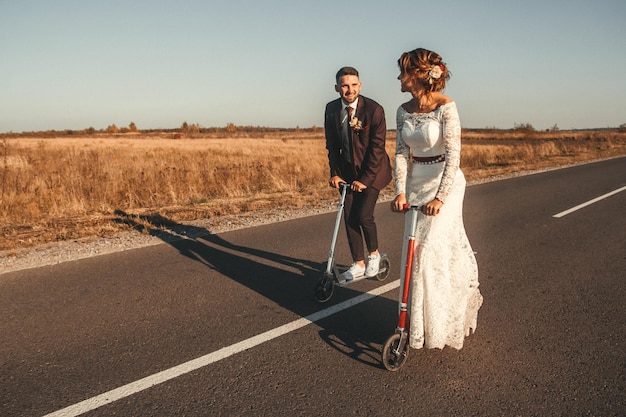 Smiling wedding couple riding a on scooters along the road outside the city at sunset