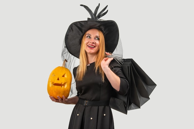 Photo smiling witch in a black costume holds a pumpkin and shopping bags exuding magic and celebration against a white studio background embodying the festive spirit of halloween