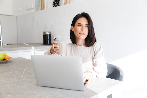  smiling woman 30s working on laptop, while sitting over white wall in bright room