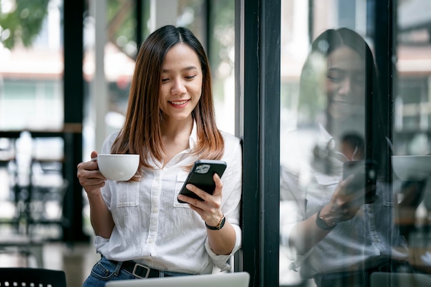 Smiling woman in casuals standing in office Businesswoman with mobile phone and cup of coffee in hand relaxing during work