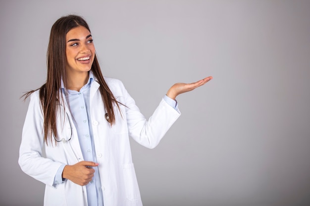 Smiling woman doctor showing aside to copyspace blank place. Confident female medical worker portrait pointing with finger to left side at empty space in studio isolated on gray background