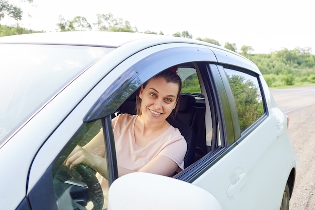 Smiling woman driver is sitting in her car, window is open, woman is looking at camera.