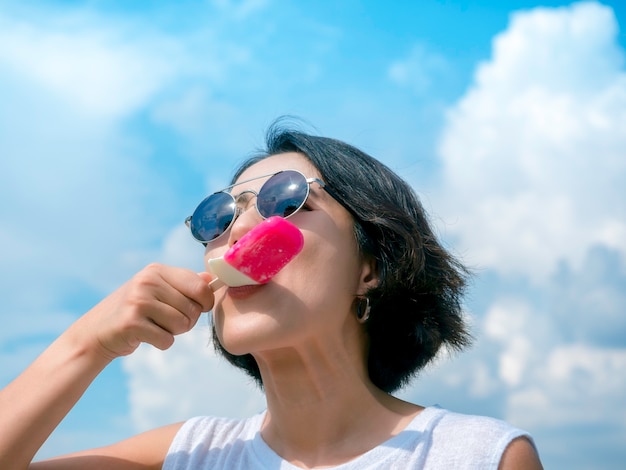 Smiling woman eating popsicles. Happy beautiful Asian woman short hair wearing sunglasses and casual white sleeveless shirt holding pink popsicle on blue sky background in summer.