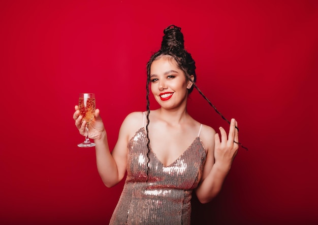 A smiling woman in an evening dress enjoys a party holding a bottle of champagne and raising a glass on a red backgrounda woman celebrates her birthday