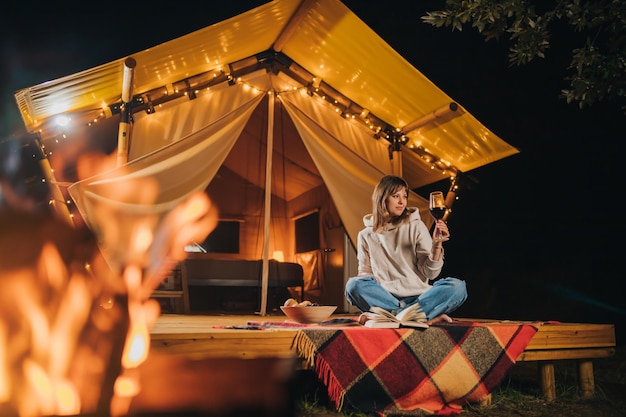 Smiling Woman freelancer drinking wine and read book sitting in cozy glamping tent in autumn evening Luxury camping tent for outdoor holiday and vacation Lifestyle concept