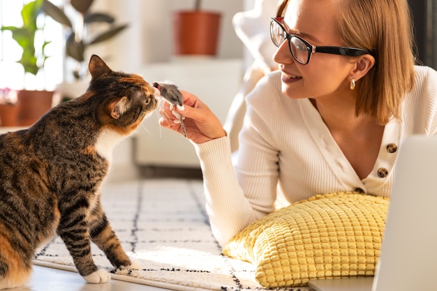 Smiling woman freelancer lies on the carpet in living room, plays with cat a toy mouse at home