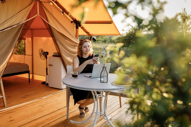 Smiling Woman freelancer using phone while working on laptop in cozy glamping tent in a sunny day Luxury camping tent for outdoor summer holiday and vacation Lifestyle concept