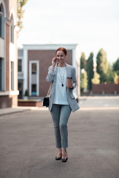 Smiling woman. Full-length portrait of a stylish young businesswoman with a paper cup of coffee smiling while talking on the phone