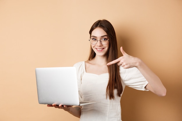 Smiling woman in glasses pointing finger at laptop screen, showing online promo, standing on beige background.